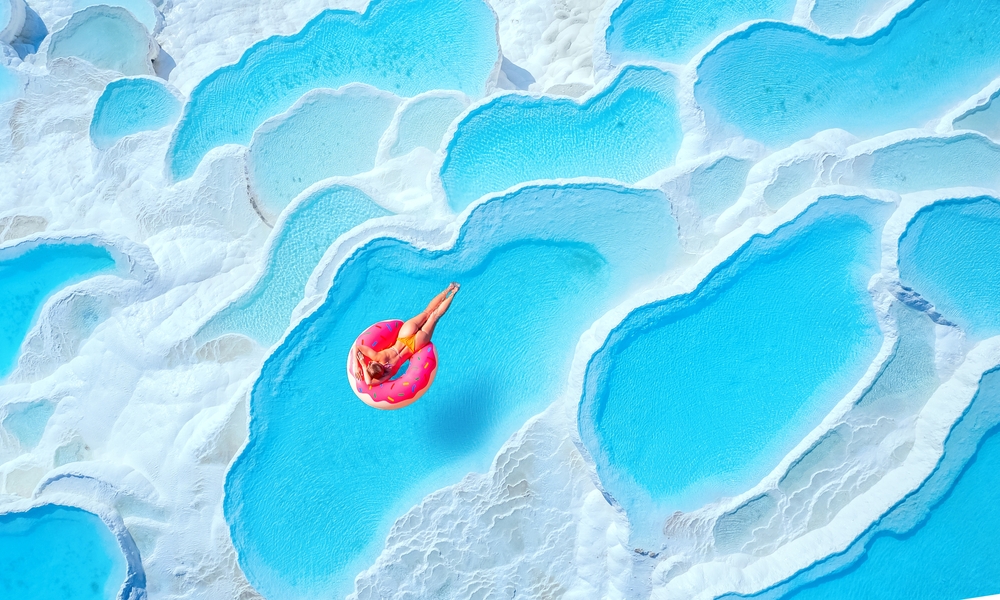 Travertine pools Pamukkale, Turkey travel. Woman swimming on pink inflatable donut in turquoise water, aerial top view.