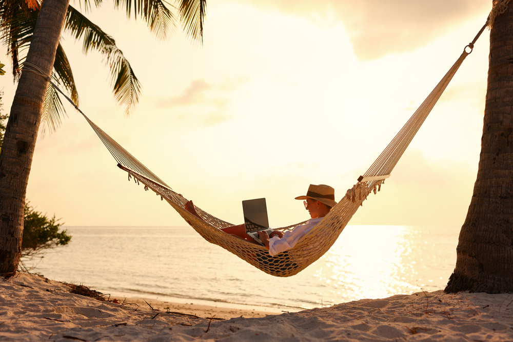 Full length of young woman, successful female freelancer using laptop while lying in hammock on the tropical beach at sunset, working remotely during summer vacation.