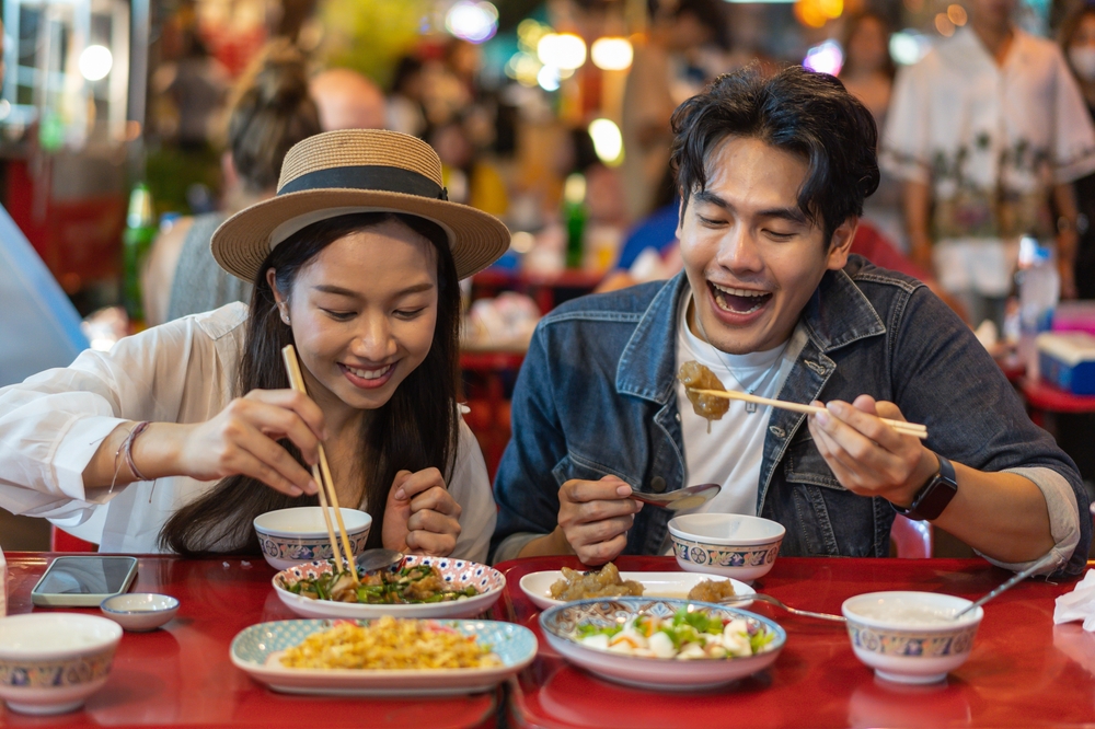 Young Asian couple traveler tourists eating Thai street food together in China town night market in Bangkok in Thailand