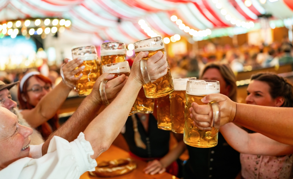 group of freinds in traditional Bavarian attire raising beer steins in a toast at a festive indoor event in an Oktoberfest or dult tent with red and white drapes in the background