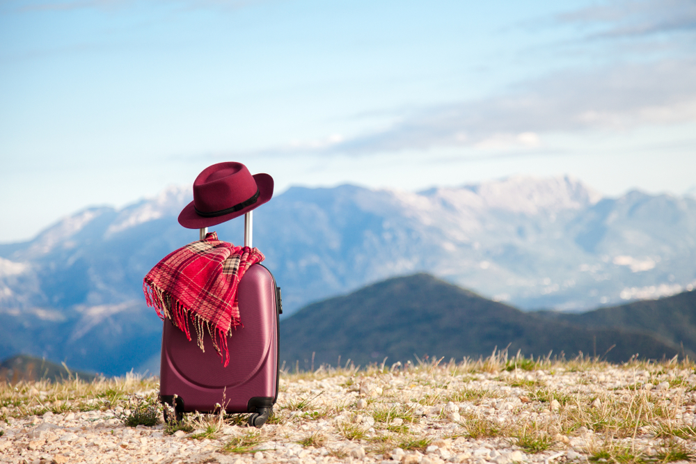 Suitcase with felt burgundy hat and plaid scarf in mountains. Concept of travel, autumn vacation, female tourism, trip, adventure.