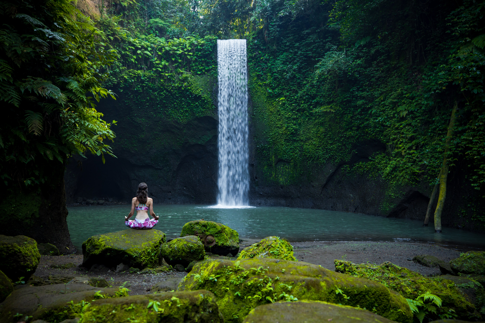 Young Caucasian woman sitting on the stone, meditating, practicing yoga, pranayama at waterfall.