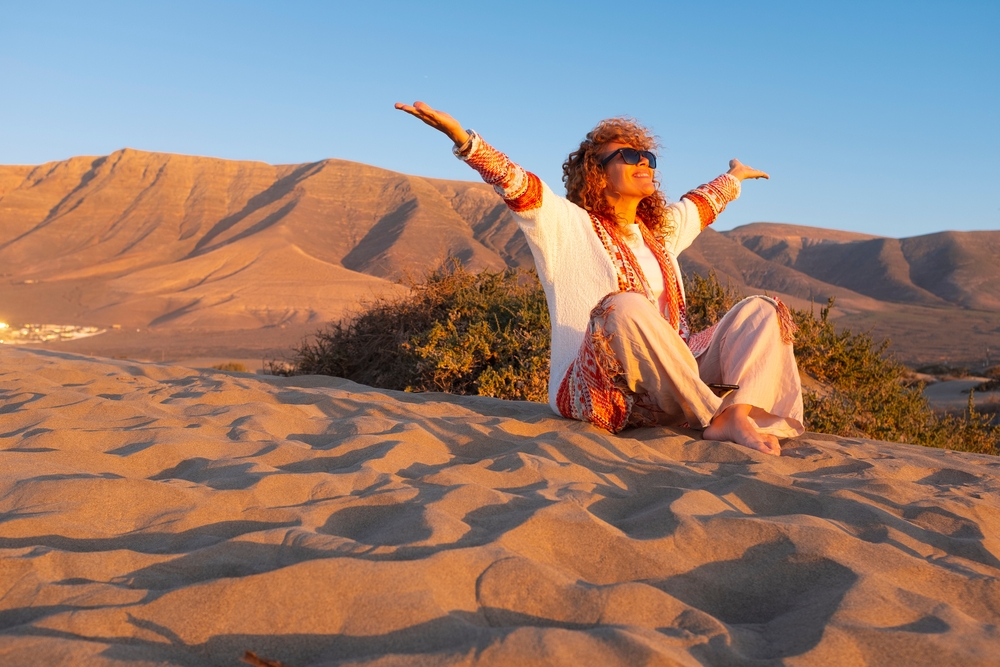 Happy people overjoyed for tourism holiday vacation. Woman sitting on the dunes sand beach outstretching arms and smile with joyful at outdoor nature around her.