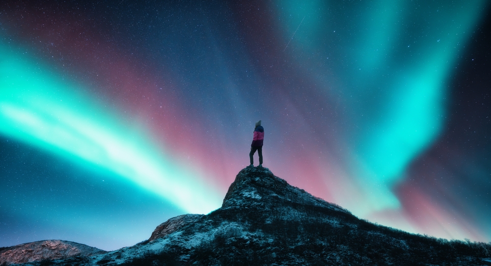 Northern lights and sporty woman on mountain peak at night. Aurora borealis and silhouette of a girl on top of rock.