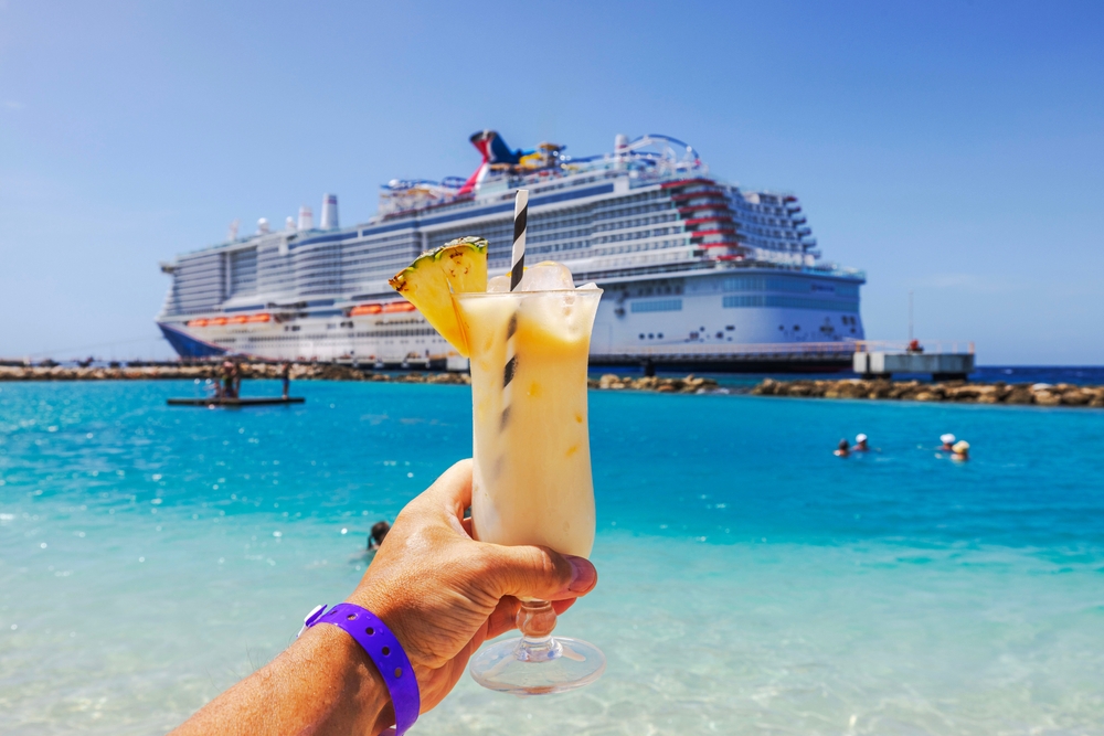 Close-up view of hand holding glass of alcoholic cocktail on Caribbean beach with cruise ship in background.