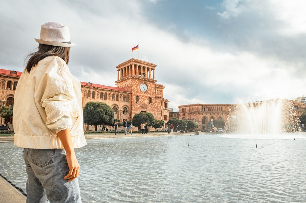 Close up woman tourist wear white jacket visit famous destination Armenia capital Yerevan. Clock bell tower with Armenian flag. Travel caucasus region in cold.Off season travel concept