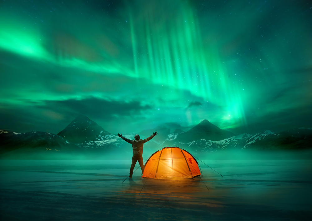 A man camping in wild northern mountains with an illuminated tent viewing a spectacular green northern lights aurora display.