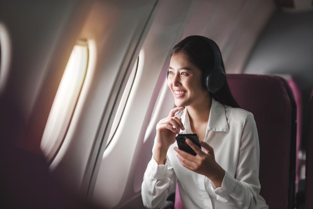 Cheerful female passenger in headphones for noise cancellation watching online movie during intercontinental flight in cabin of aircraft, happy young woman using wifi connection on board