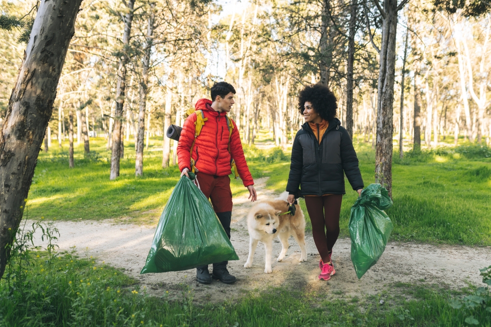 Young couple cleaning up litter in a forest with their dog, carrying large green bags and contributing to environmental conservation.