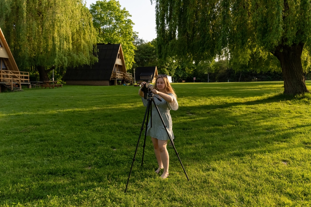 Female photographer with camera on tripod amidst green landscape and wooden houses. Holiday and relaxation in a natural environment. High quality photo