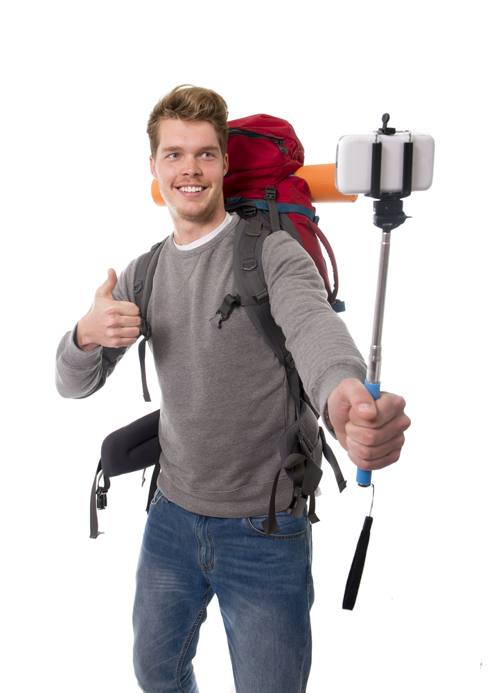 young attractive backpacker tourist taking selfie photo with stick carrying backpack ready for travel and adventure on vacations route isolated on white background