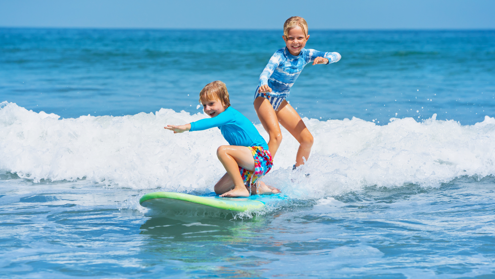 Happy baby boy and girl - young surfers ride with fun on one surfboard.