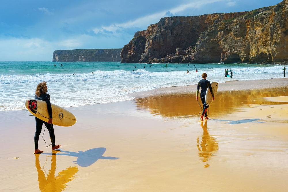 Young men walking by sandy beach with surfboard. ALgarve is a famous surfing destination in Portugal