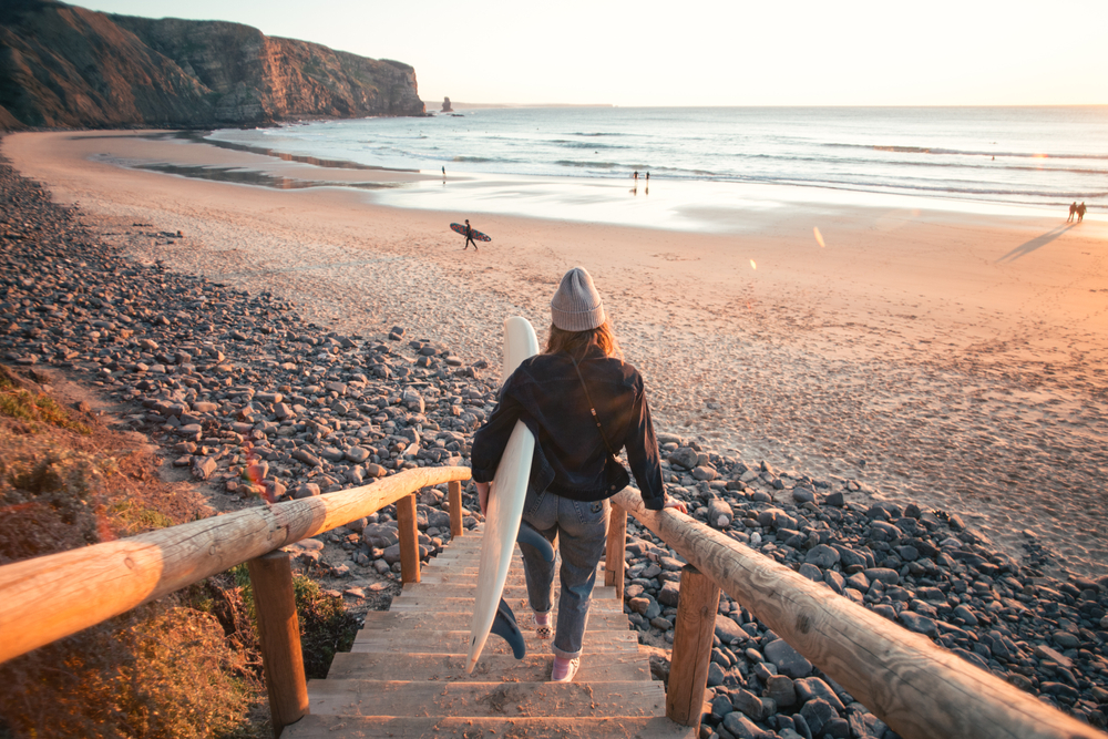 Young casual dressed woman walk down wooden stairs or walk board to epic beach at sunset, carry professional surfboard ready for evening surf session. 
