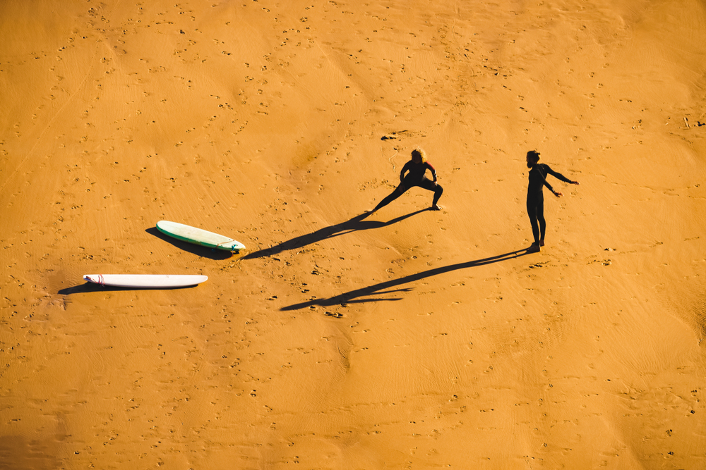 Young adults surfers wearing wetsuit warming up on beach preparing for summer evening surfing session - stretching routine for surfing.