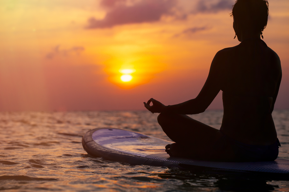 Woman practicing SUP yoga at sunset, meditating on a paddle board.