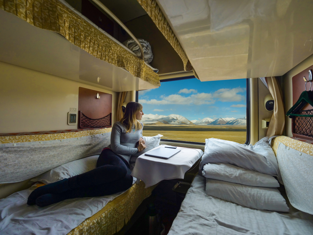 Young woman lies on the bed and looks through the window of the sleeper train crossing picturesque Tibet.