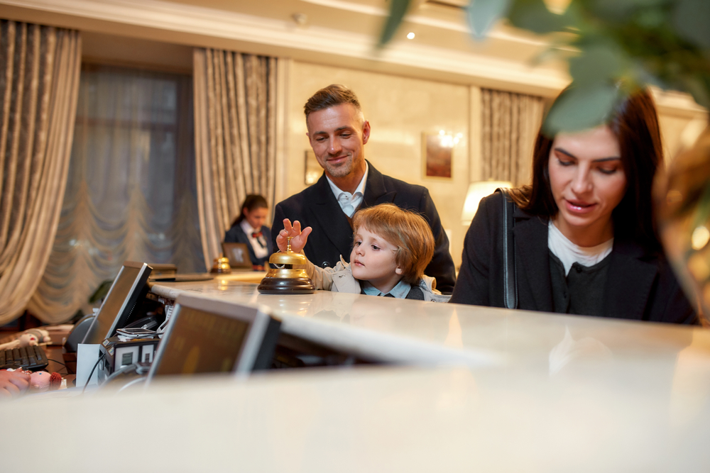 Happy family checking in hotel at reception desk. A son is ringing a service bell while standing with parents at the hotel reception