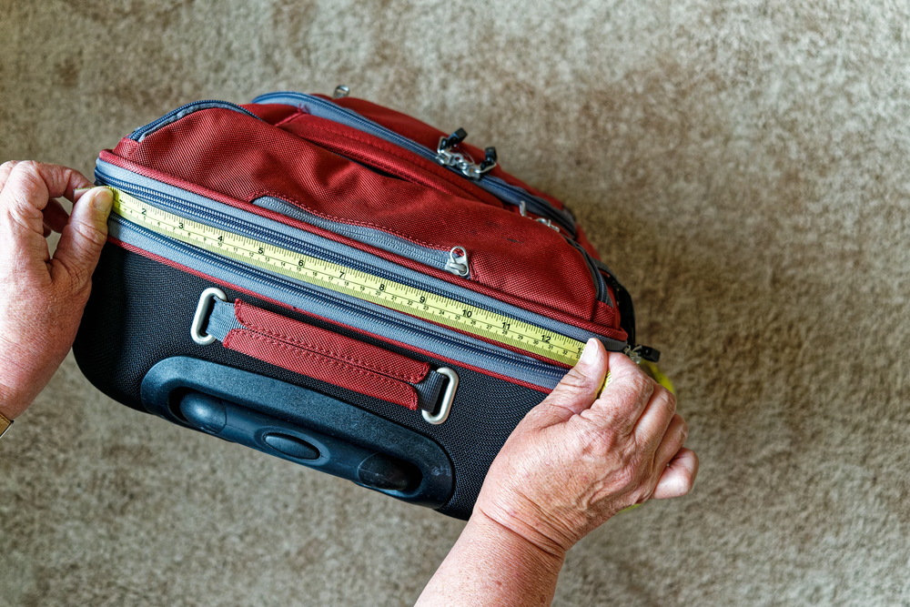 caucasian senior citizen female hands with yellow tape measure with inches and centimeters measuring a red carry on size piece of luggage at home viewed from above