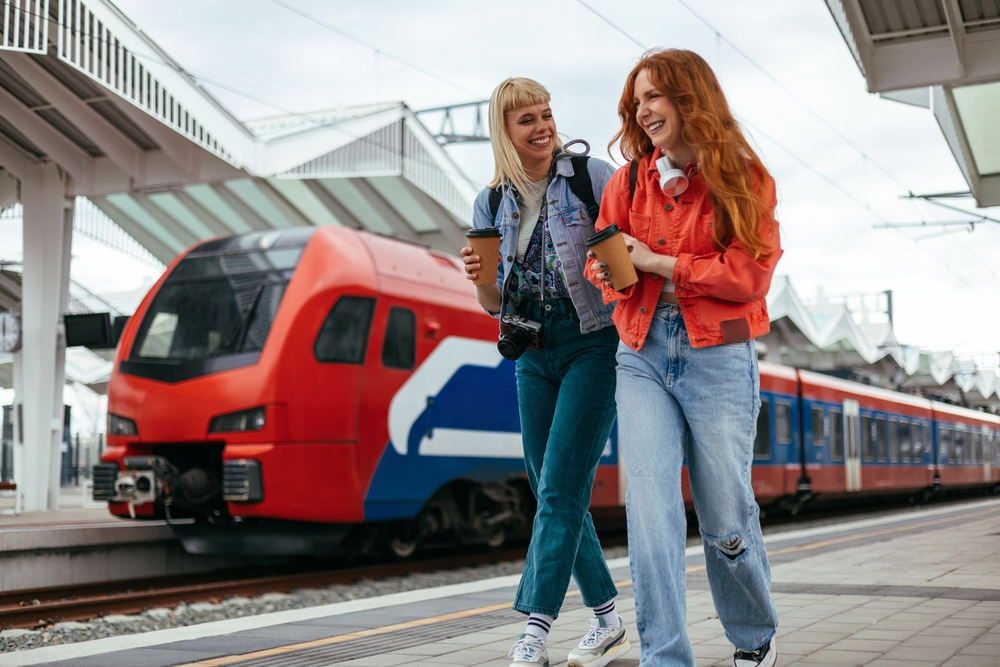 Two young women walking together at a train station with luggage, with a red train in the background