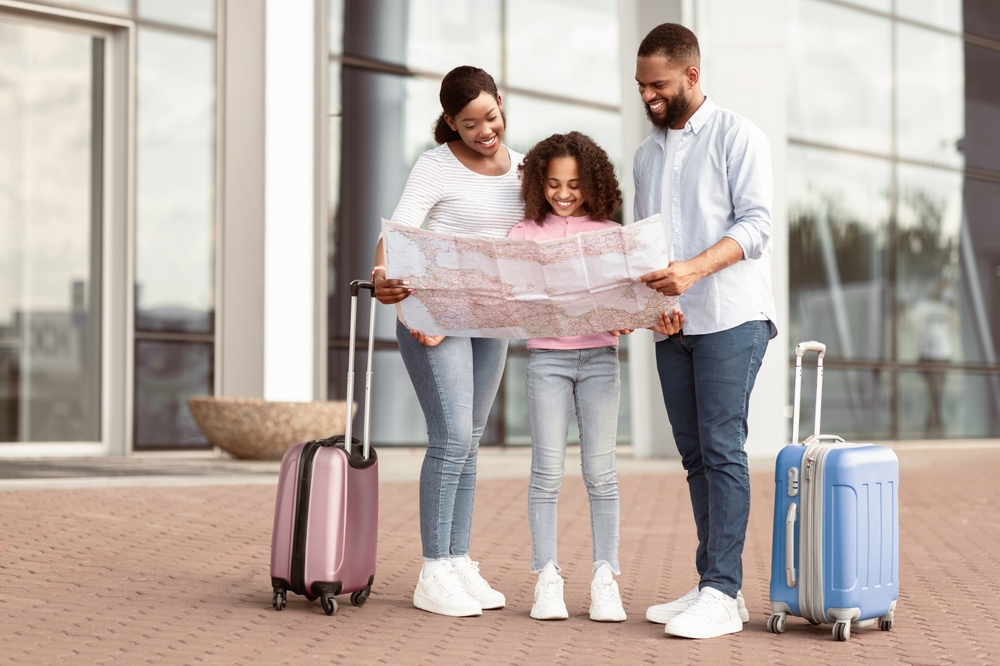 Happy Black Tourists Standing Near Airport, Checking City Map, Cheerful African American Man, Woman And Girl Planning Trip Route.