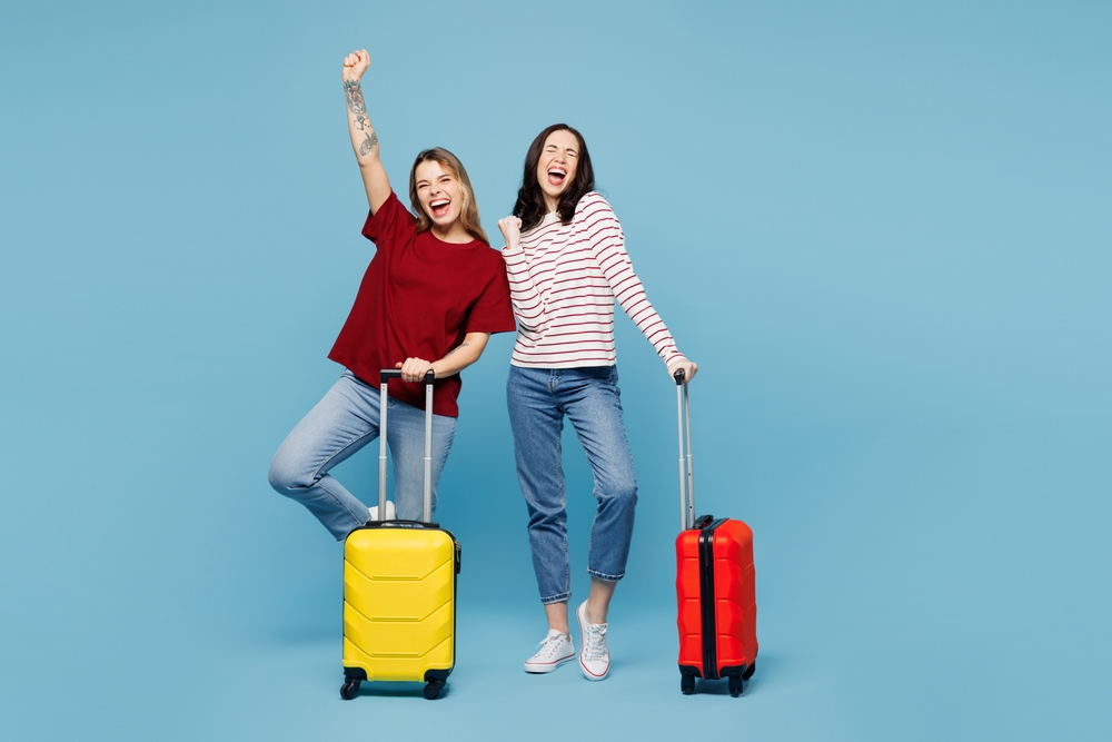 Traveler couple two friends women wear red casual clothes hold bag do winner gesture isolated on plain blue background.