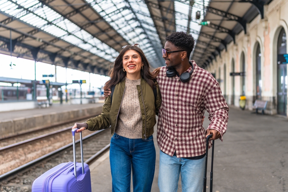 Young multi ethnic couple walking together with luggage at a train station platform, enjoying their vacation