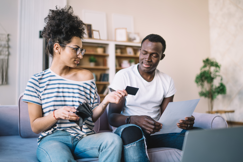 Ethnic female sitting on sofa at home and giving African American husband credit card for paying household bills by laptop