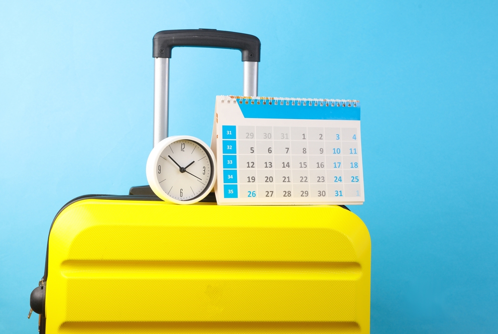 Vacation planning. Yellow travel suitcase with calendar, clock on a blue background.