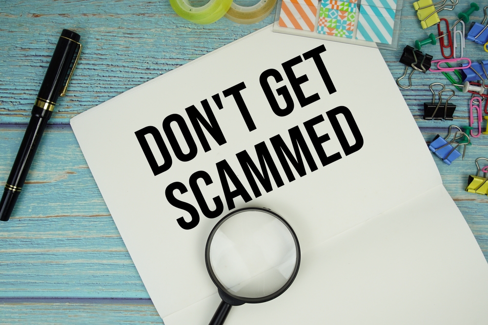 Don't Get Scammed text on a paper surrounded by office supplies and magnifying glass on a blue wooden surface